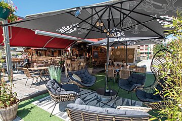 A sunny outdoor cafe patio featuring black umbrellas, red awnings, wicker and wooden seating areas, lush plants, and a mix of artificial turf and wooden decking.