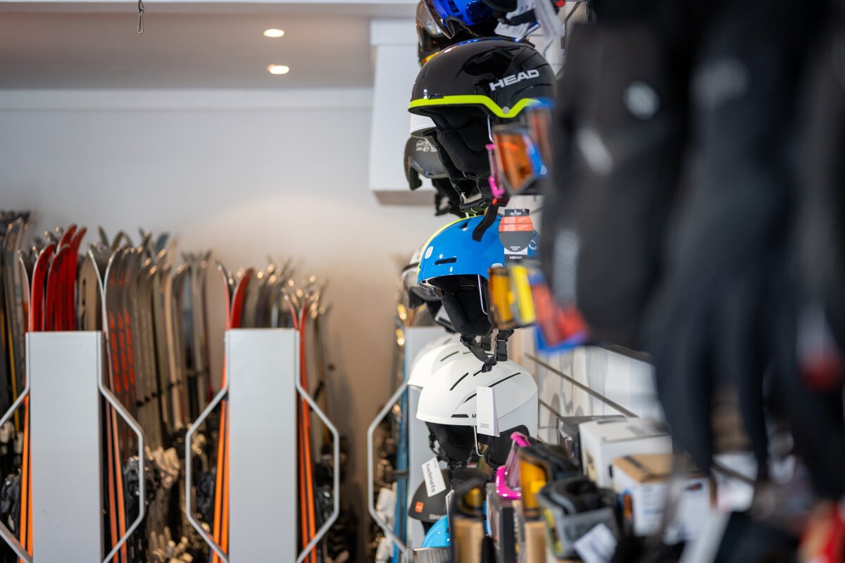 A ski shop interior displays rows of colorful helmets and goggles on the right, with racks of skis visible in the background on the left.