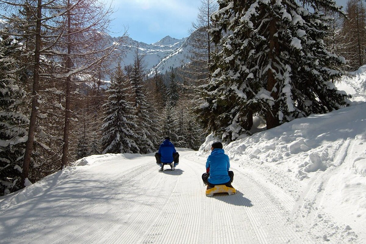 Two people sledding down a snowy road with mountains in the background