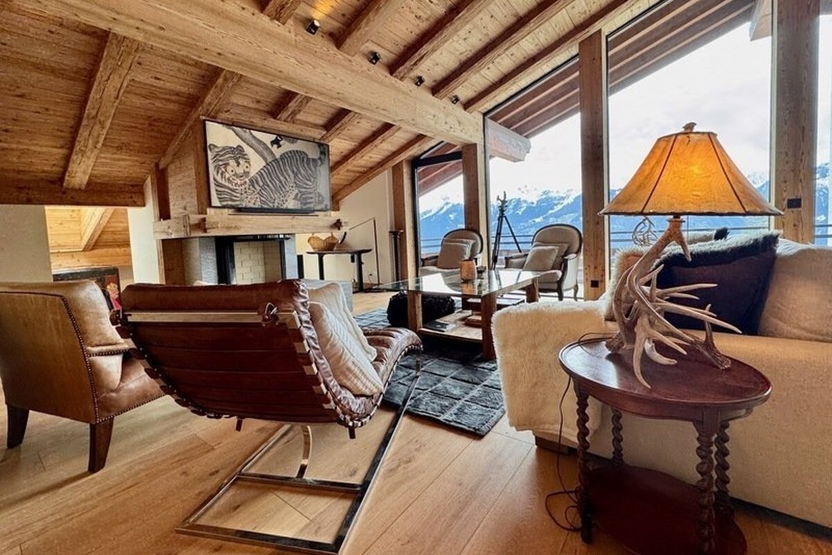 Cozy rustic-modern chalet living room with vaulted wood ceilings, leather chairs, and large windows overlooking snow-capped mountains.