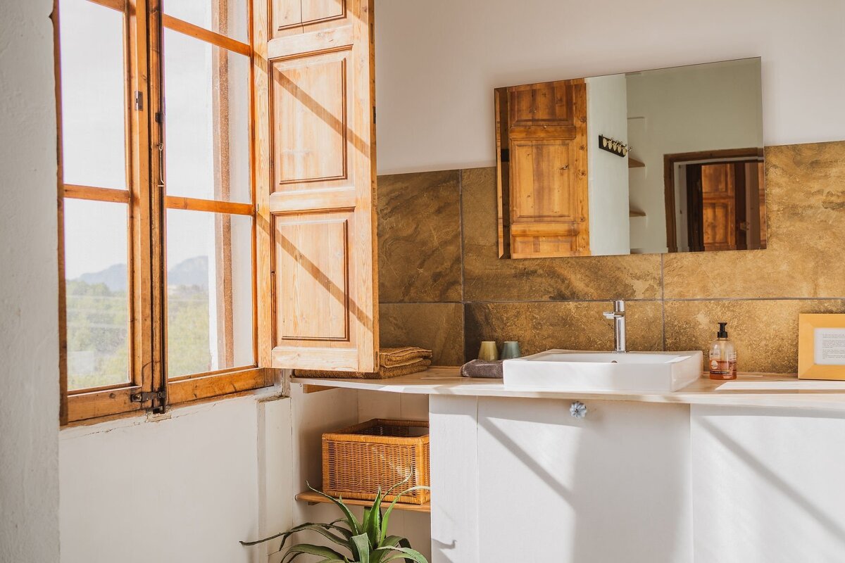 A sunny, rustic bathroom features open wooden shutters, a modern square sink on a vanity, and a mirror, all bathed in warm sunlight.