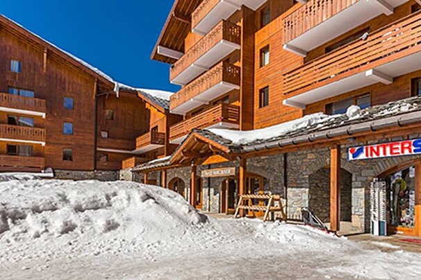 A sunny, snowy ski resort features multi-story wooden chalet-style buildings with balconies, snow on roofs, and an 