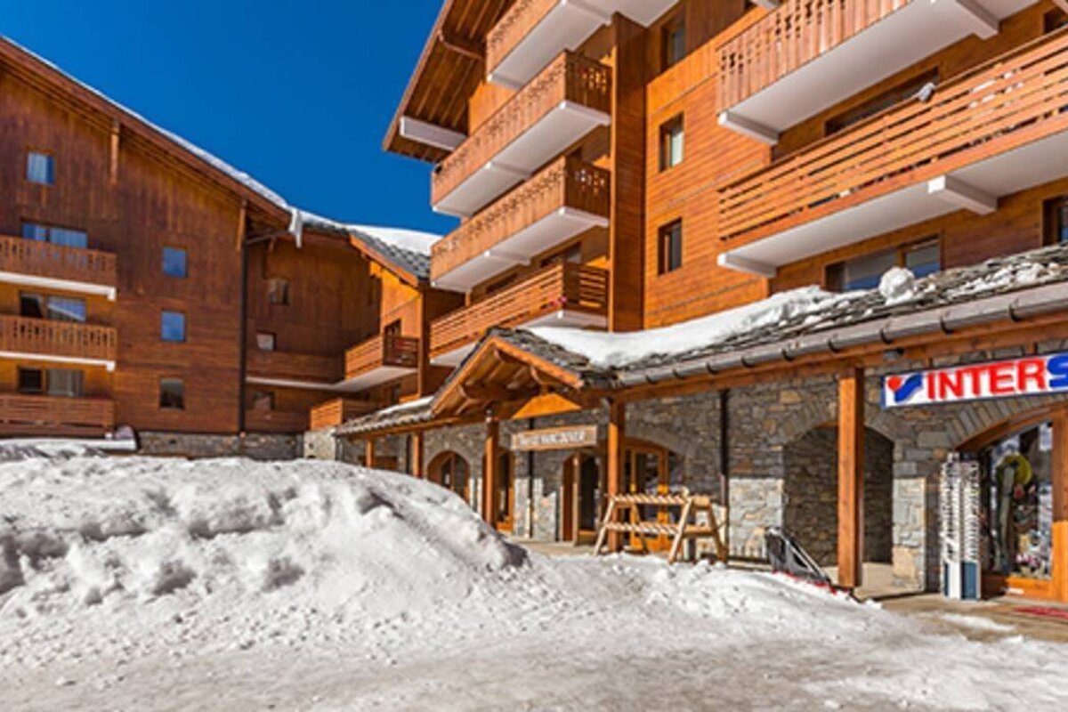 A sunny, snowy ski resort features multi-story wooden chalet-style buildings with balconies, snow on roofs, and an