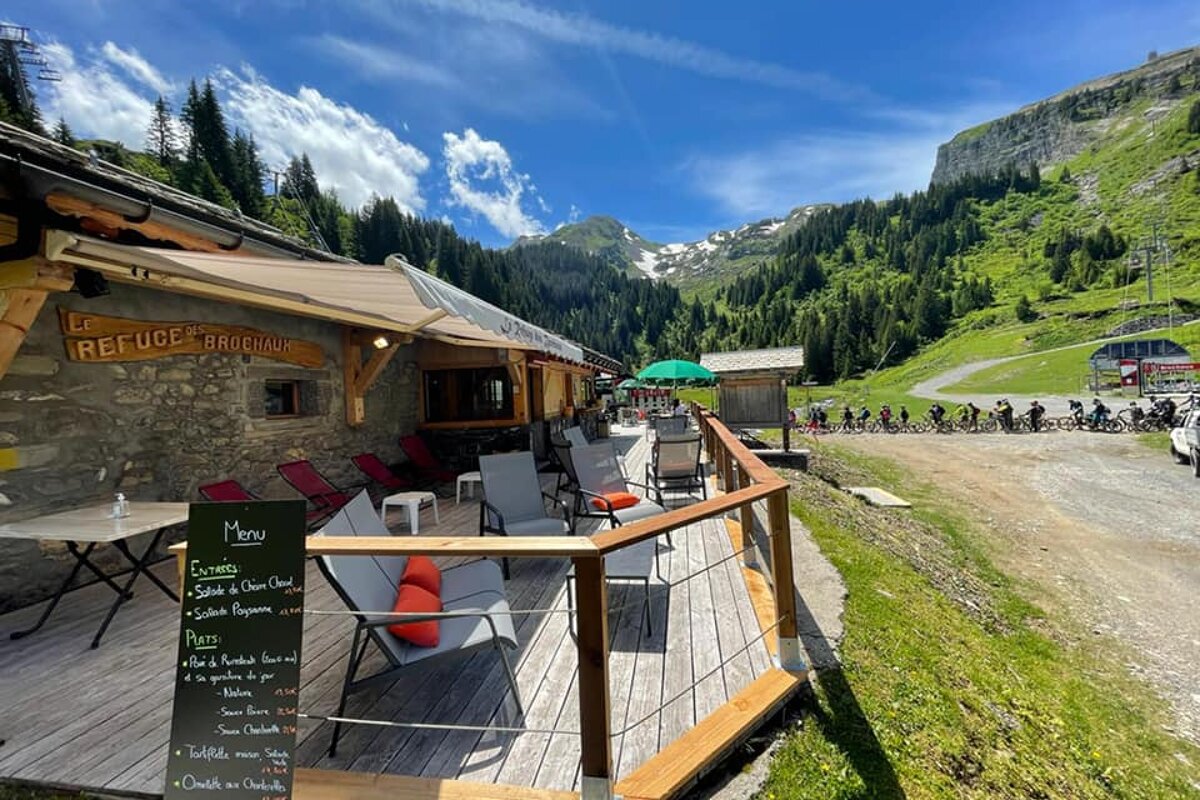 A mountain refuge with a sunny deck overlooks a scenic valley. A line of mountain bikers pauses on a dirt path, surrounded by green slopes and snowy peaks.