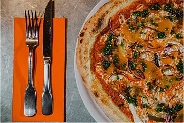 A vibrant pizza with pesto, bell peppers, and red onions, served on a white plate next to a fork and knife on an orange napkin.