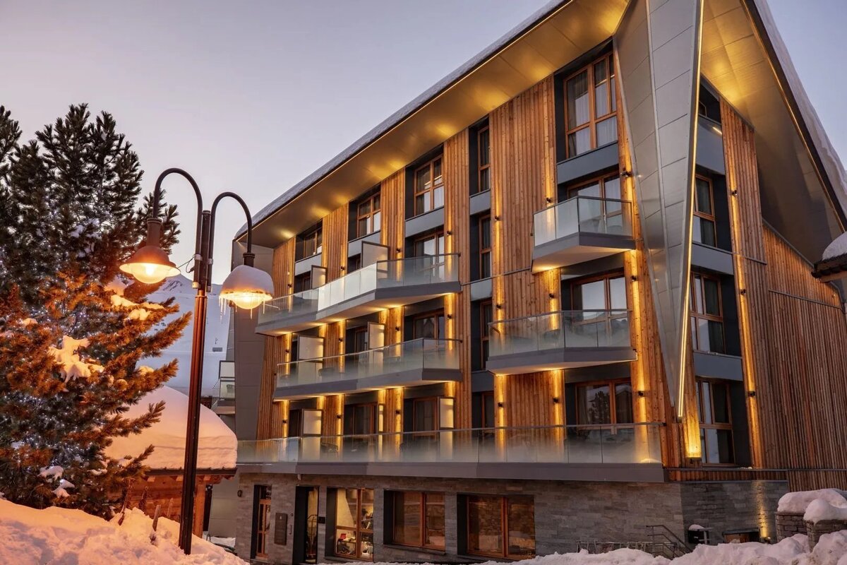 A modern wood and glass hotel, warmly lit at dusk, stands amidst snow. A streetlamp with glowing bulbs and icicles is in the foreground beside a snowy pine.