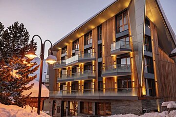 A modern wood and glass hotel, warmly lit at dusk, stands amidst snow. A streetlamp with glowing bulbs and icicles is in the foreground beside a snowy pine.