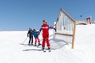 A ski instructor teaches two children on a sunny mountain slope, with a large outdoor chimes structure and a