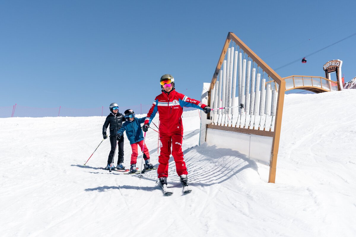 A ski instructor teaches two children on a sunny, snowy mountain slope, passing a large chime structure and the