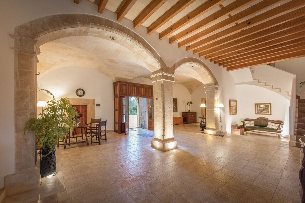 A grand, elegant hall featuring stone arches, pillars, vaulted ceilings, and wooden beams. Tiled floors, classic furniture, and an open doorway lead to the exterior.