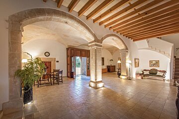 A grand, elegant hall featuring stone arches, pillars, vaulted ceilings, and wooden beams. Tiled floors, classic furniture, and an open doorway lead to the exterior.