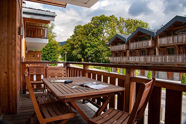 A rustic wooden balcony with a table and chairs overlooks green trees and chalet-style buildings with dark roofs, under a cloudy sky.