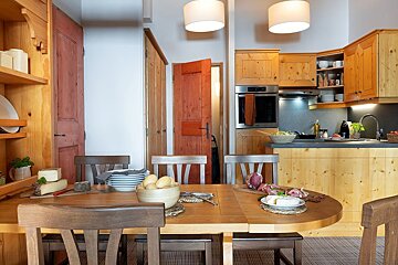 A cozy, rustic dining area with a wooden table set with food, leading into a kitchen with light wood cabinets.