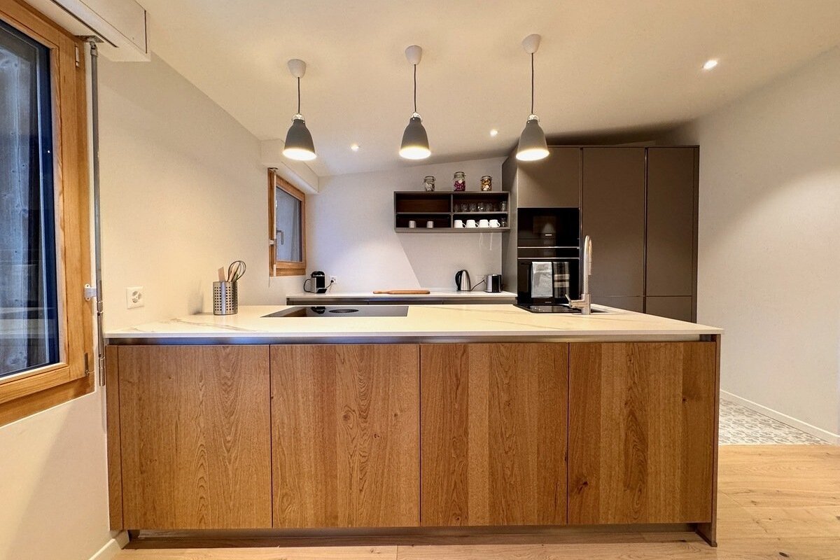 A modern kitchen with a large wooden island, white countertop, gray pendant lights, and dark gray built-in appliances and cabinets.