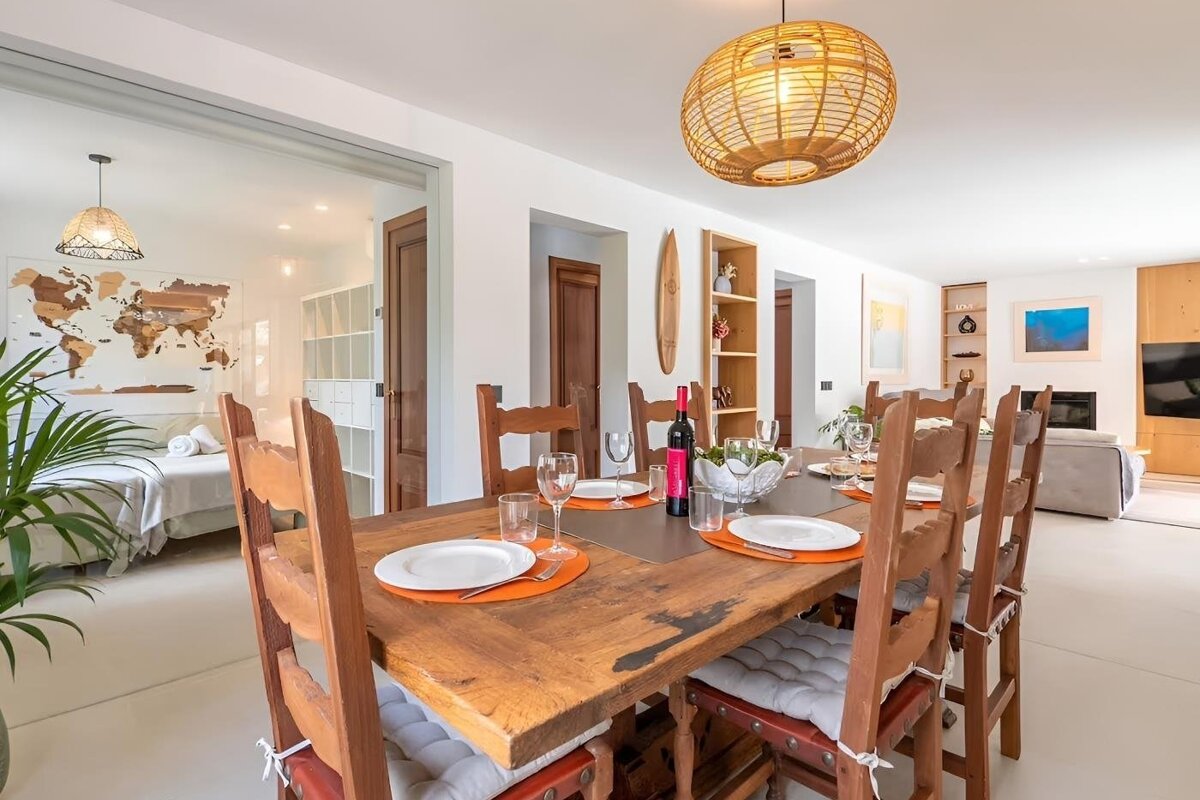A bright, open-plan dining room with a rustic wooden table set for dinner, a rattan lamp, and views of a stylish living area and bedroom.