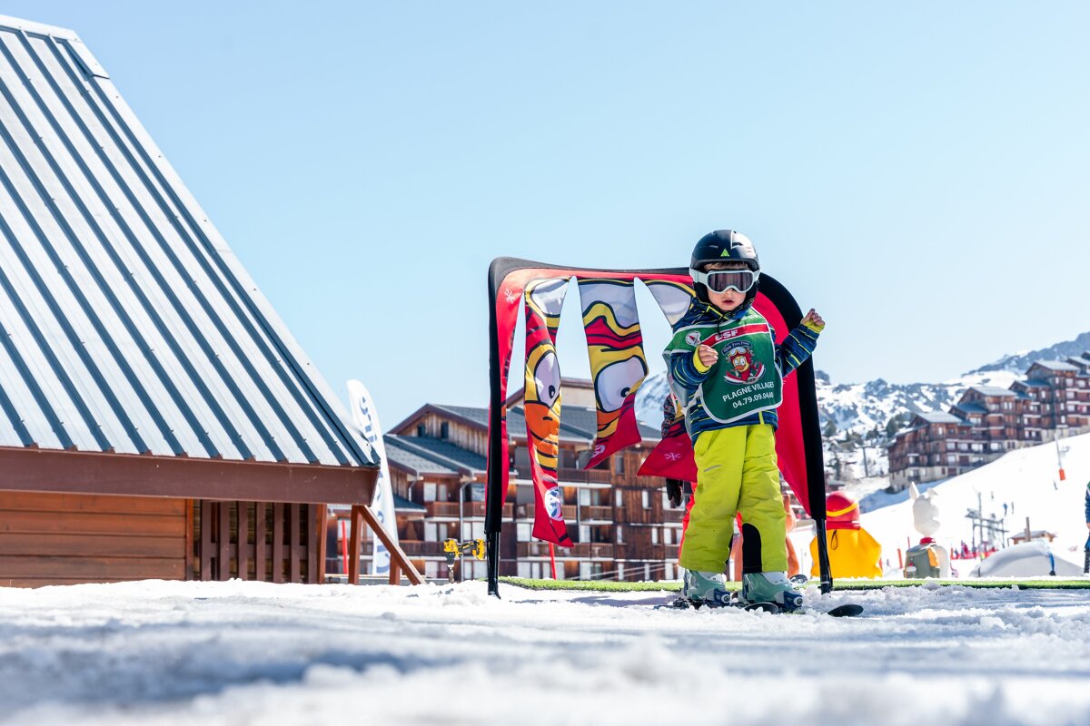 A young child in ski gear (helmet, goggles, green bib) stands on snow at a sunny ski resort, with colorful flags and buildings behind them.