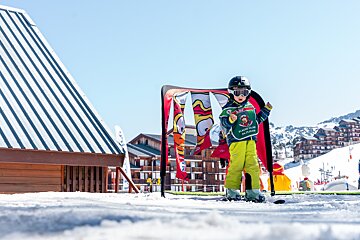 A young child in ski gear (helmet, goggles, green bib) stands on snow at a sunny ski resort, with colorful flags and buildings behind them.