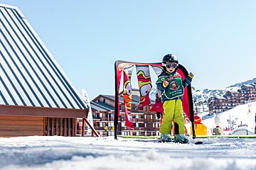 A young skier in full gear (helmet, goggles, green bib) stands on skis in a sunny snowy mountain resort with buildings and colorful banners.