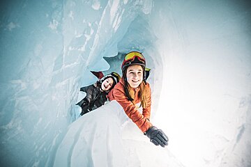 Two smiling girls, wearing helmets and winter jackets, peek out from a luminous blue ice cave, their faces showing pure joy.