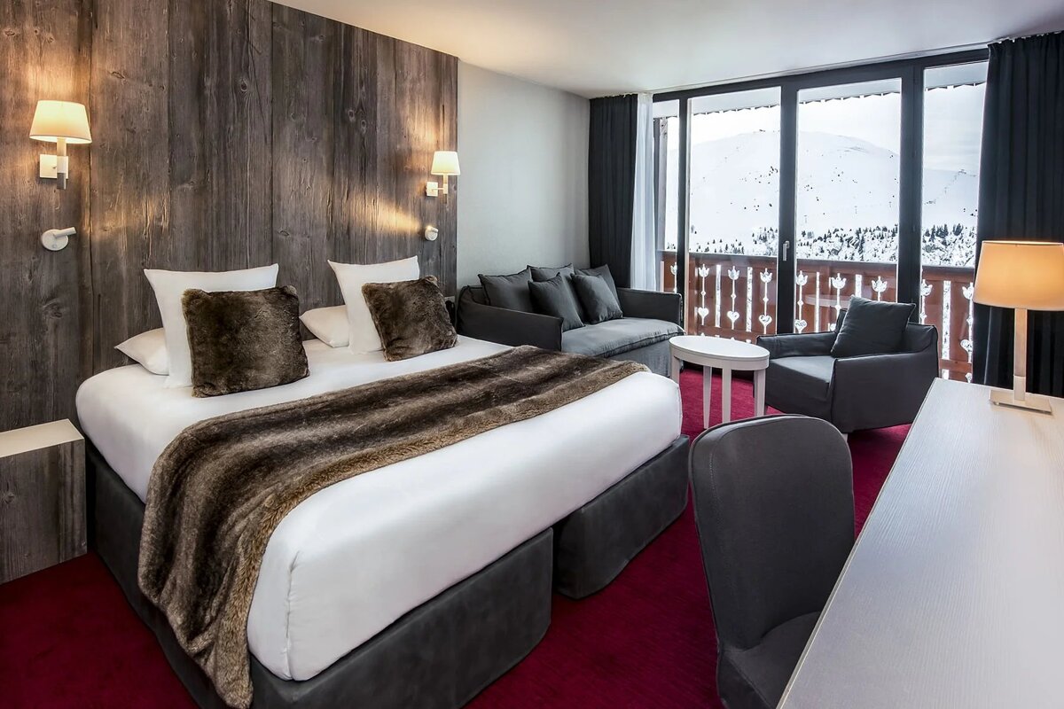 A cozy mountain hotel room with a bed, sofa, desk, and balcony overlooking snowy peaks. Features rustic wood paneling, a faux fur throw, and red carpet.