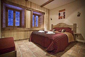 Cozy rustic bedroom features wooden walls, exposed beams, a large bed with red bedding, tiled floor, and windows revealing a snowy forest scene outside.
