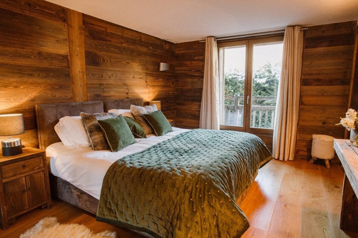 Cozy rustic bedroom with rich wooden walls, a large bed featuring green bedding, and a balcony window overlooking lush trees, warmly lit.