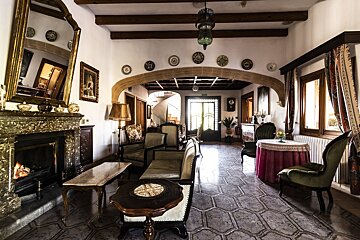 An elegant, traditional lounge with a grand fireplace, ornate mirror, plush seating, tiled floors, and wooden beams, exuding an antique charm.