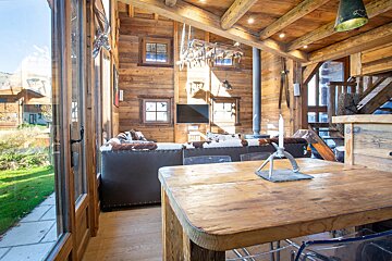 A rustic wooden chalet interior with a living room, dining table, and large windows revealing a mountain view. Lodge decor and a cozy fireplace are visible.