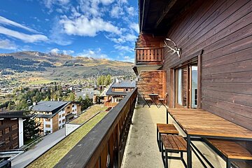 A long wooden balcony with outdoor seating overlooks a sprawling mountain village and valley under a bright blue, cloudy sky. Antlers adorn the chalet wall.