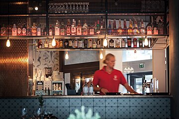 A man in a red shirt stands behind a bar with many bottles of liquor on it