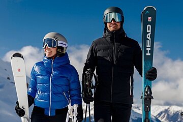 A man and a woman in ski gear smile while holding Head skis against a bright blue sky and snowy mountains.