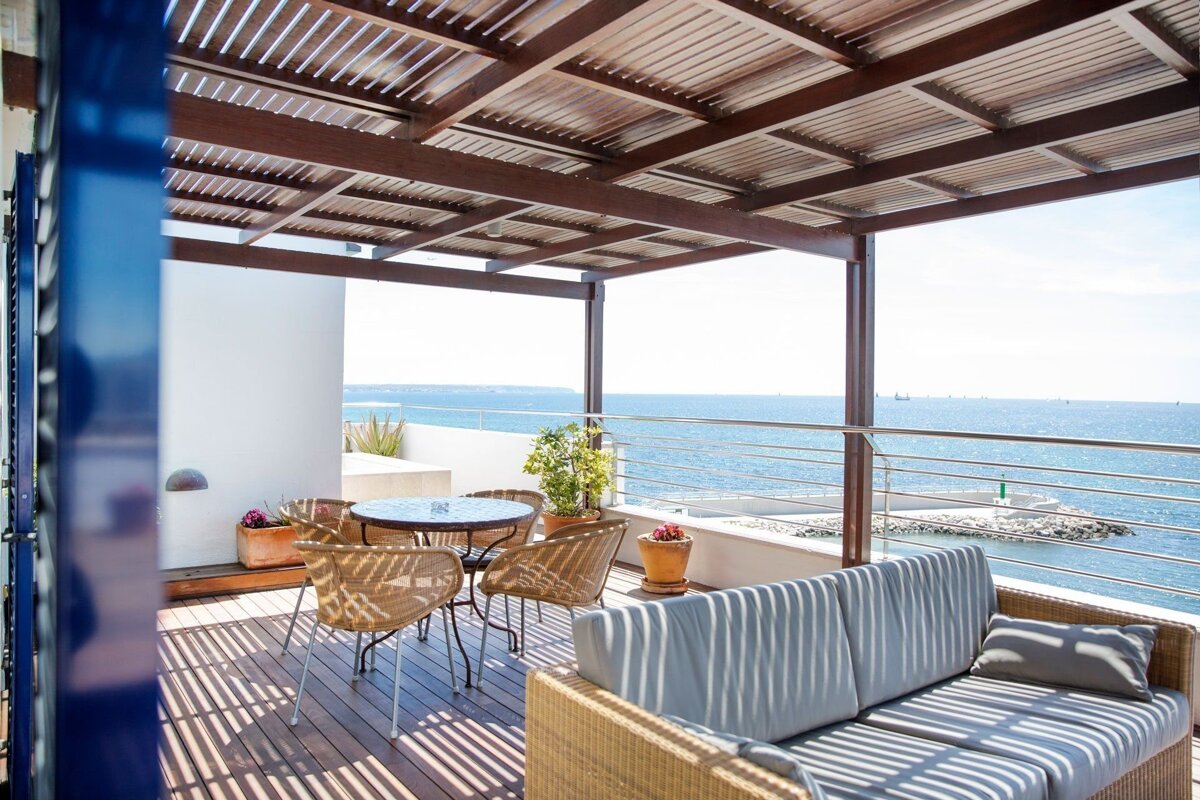Sunny outdoor patio with a pergola, lounge furniture, and dining set, overlooking a bright blue sea. Sunlight casts striped shadows on the wooden deck.