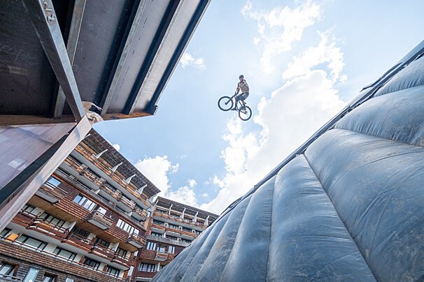 A biker performs an aerial trick against a blue sky, above a large inflatable bag, with multi-story buildings framing the scene.