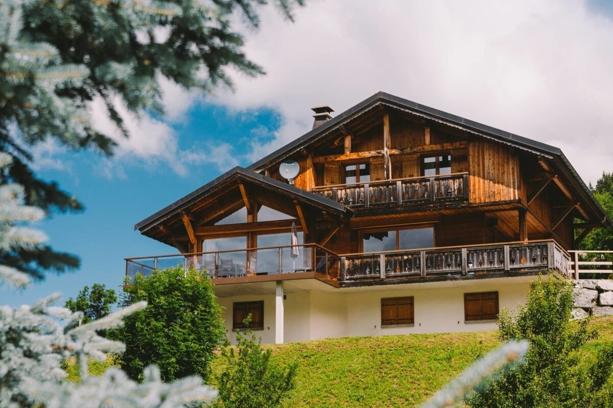 A cozy wooden chalet-style house with multiple balconies nestled on a lush green hill under a partly cloudy blue sky, framed by blurred pine branches.
