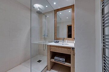 A modern bathroom with a glass-enclosed shower, wooden vanity, framed mirror, and white sink. Light tiled walls and a chrome towel warmer are visible.