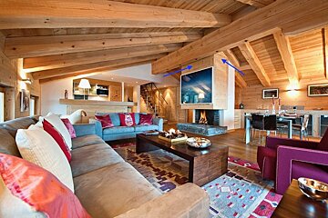 A cozy, luxurious chalet-style living room with beamed wooden ceilings, a fireplace under a TV, comfy sofas, and a dining area, exuding warmth.