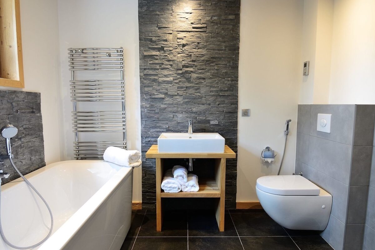 A modern bathroom with a white tub, toilet, and vessel sink on a wooden stand. A dark stone accent wall contrasts with light walls and chrome fixtures.