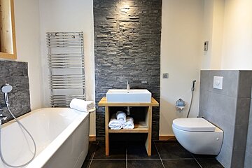 A modern bathroom with a white tub, toilet, and vessel sink on a wooden stand. A dark stone accent wall contrasts with light walls and chrome fixtures.