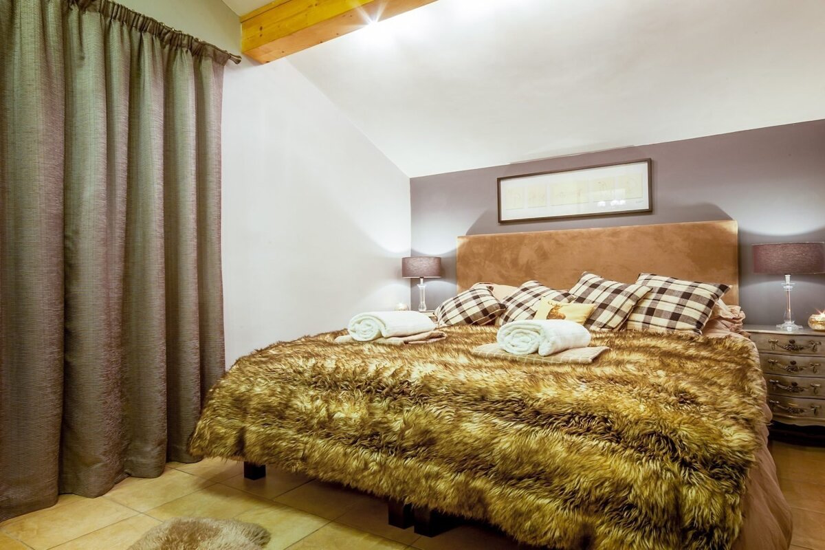 A cozy bedroom featuring a bed with a brown faux fur throw, plaid pillows, and bedside lamps. Neutral walls and dark curtains complete the warm space.