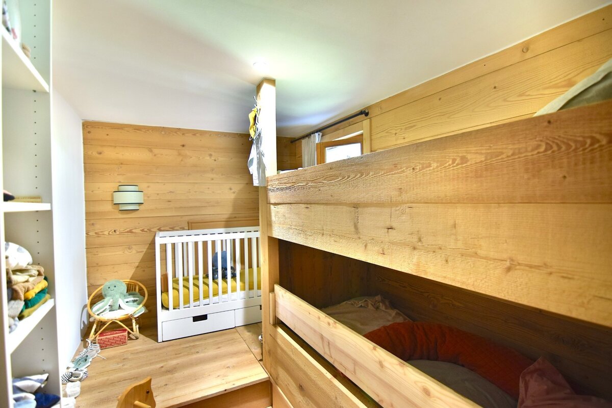 A cozy kids' room with wood-paneled walls, built-in wooden bunk beds, a white crib, a rattan chair, and a shelving unit. Bright and inviting.
