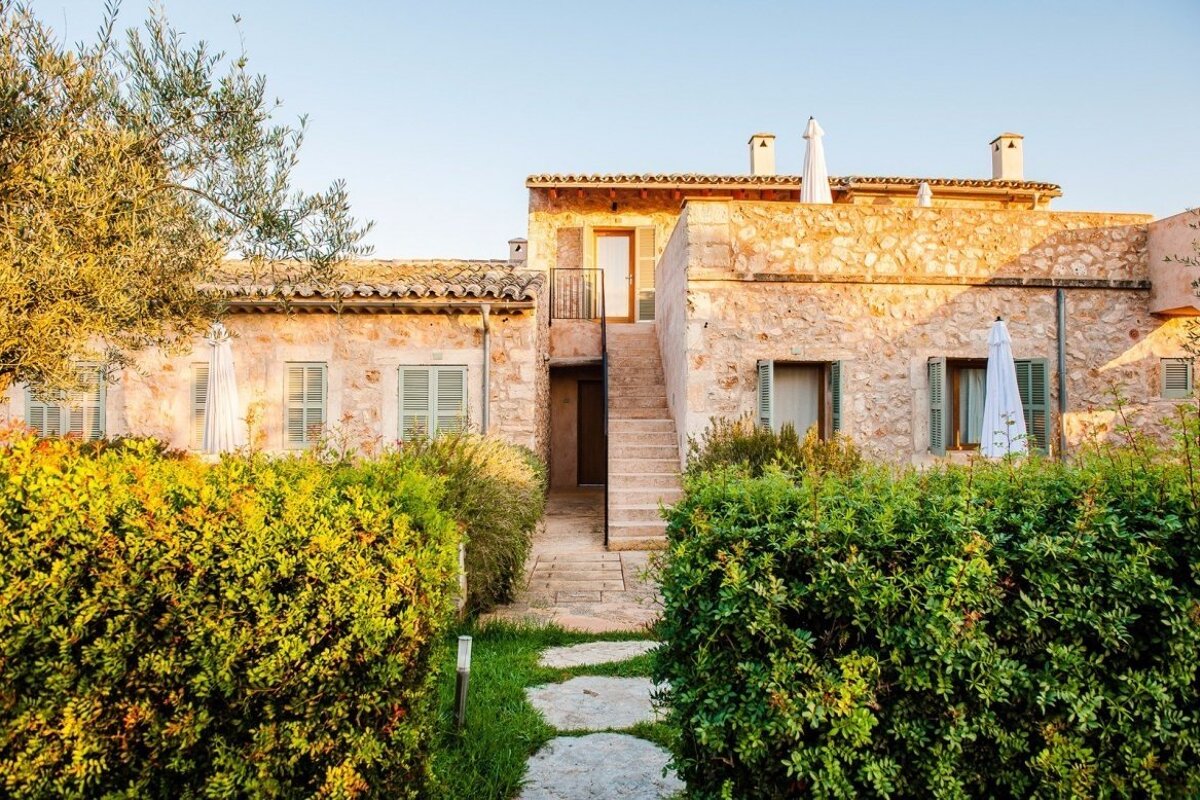 Sunlit stone building with green shutters, tiled roof, and an external staircase. Lush hedges frame a stepping stone path under a clear blue sky.