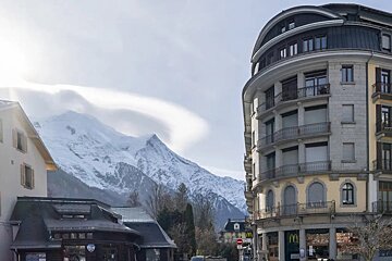 Snowy mountains dominate the background of a town with buildings, including a curved one housing a McDonald's. Lenticular clouds hover above the peaks.