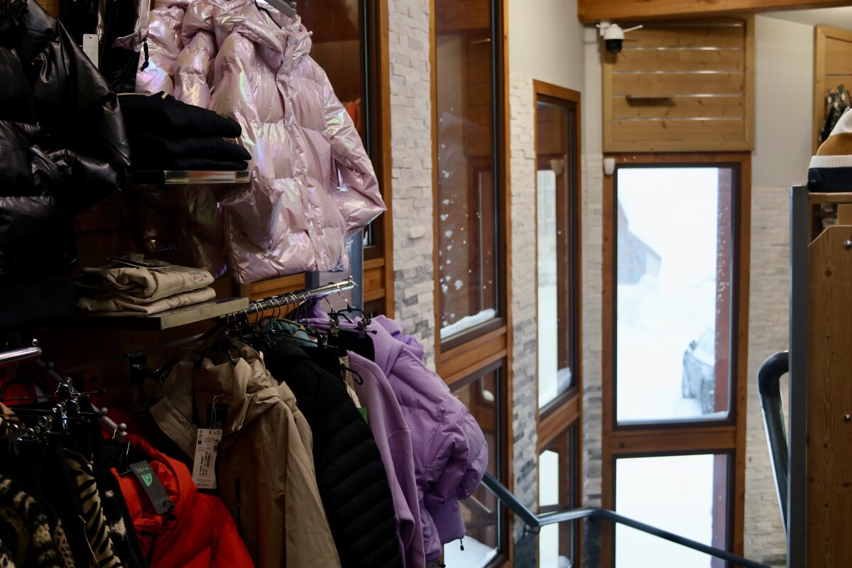 A shop displays various winter jackets, prominently a shiny pink puffer, while large windows reveal a snow-covered street and car outside.