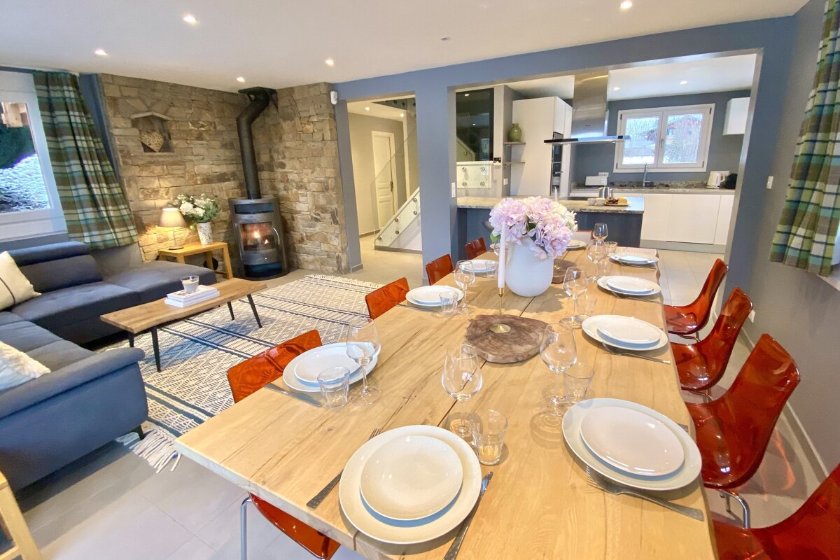 An open-plan living space with a large wooden dining table, orange chairs, blue sectional sofa, a stone wall with a wood stove, and a modern kitchen.