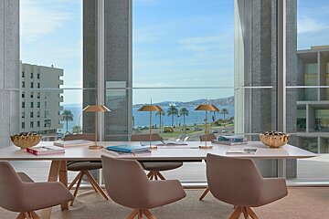 Modern meeting room with a white table and chairs, offering a panoramic view of the blue ocean, palm trees, and a coastal road through large windows.