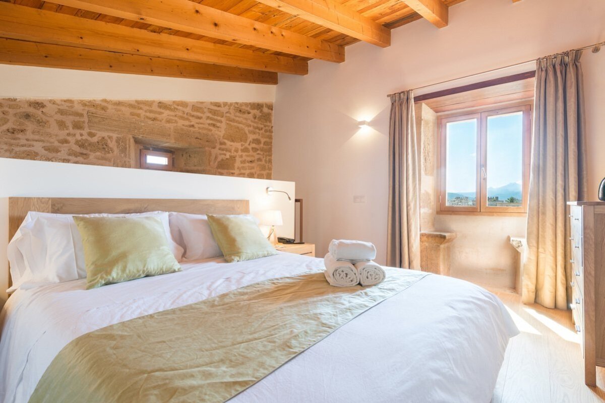 Bright bedroom with a large bed, exposed wooden beams, a stone wall, and a window overlooking a sunny landscape.