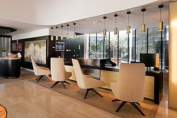 Modern hotel lobby with a sleek reception desk, beige chairs, and gold pendant lights. Large windows with blinds offer natural light.