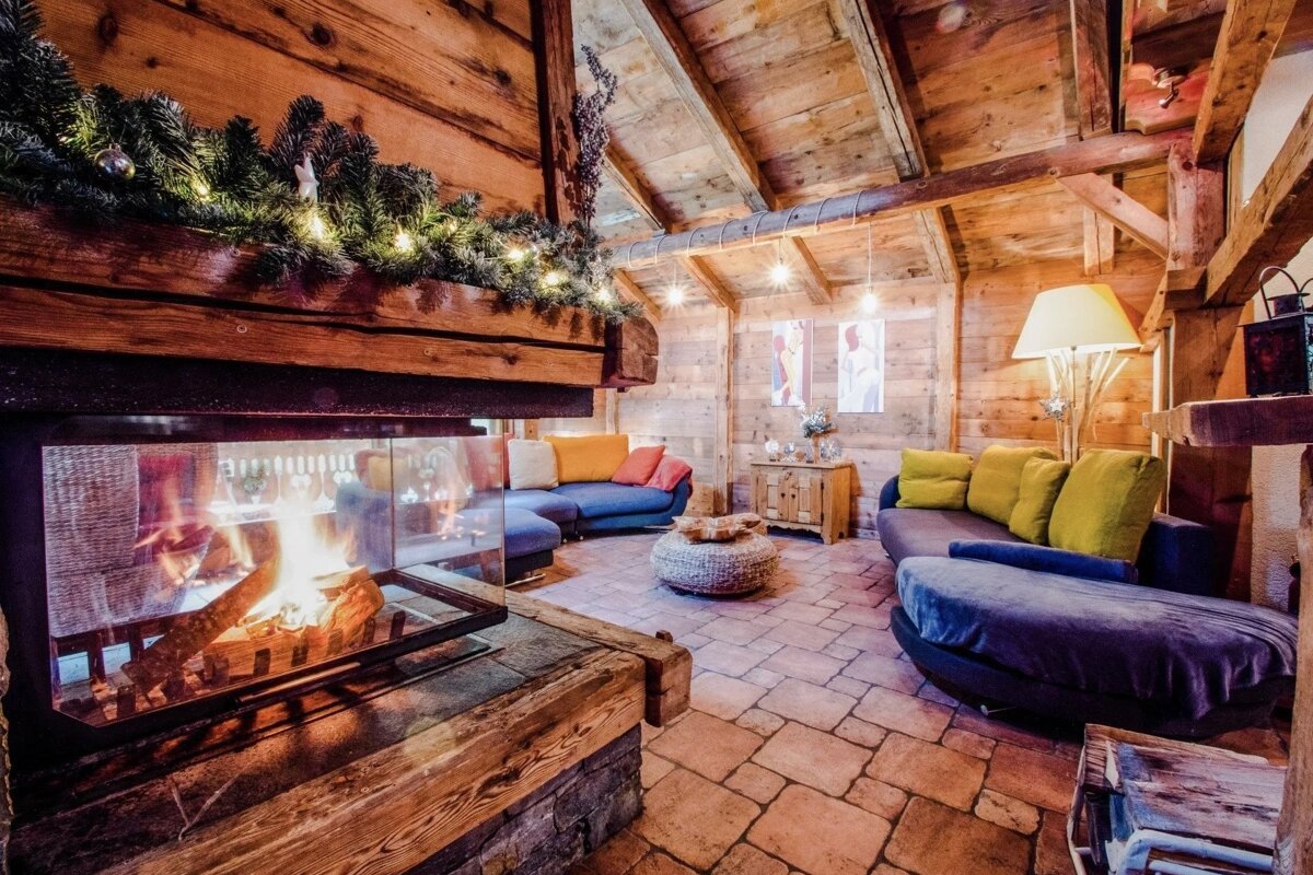 A warm, rustic wooden room with a crackling fireplace, blue/yellow sofas, and festive garland. Cozy chalet ambiance.