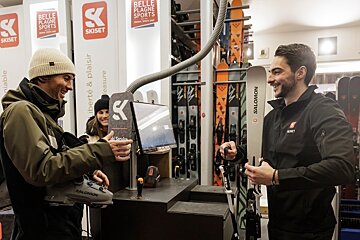 A ski shop employee helps a customer holding ski gear. Indoor shop, racks of skis behind.