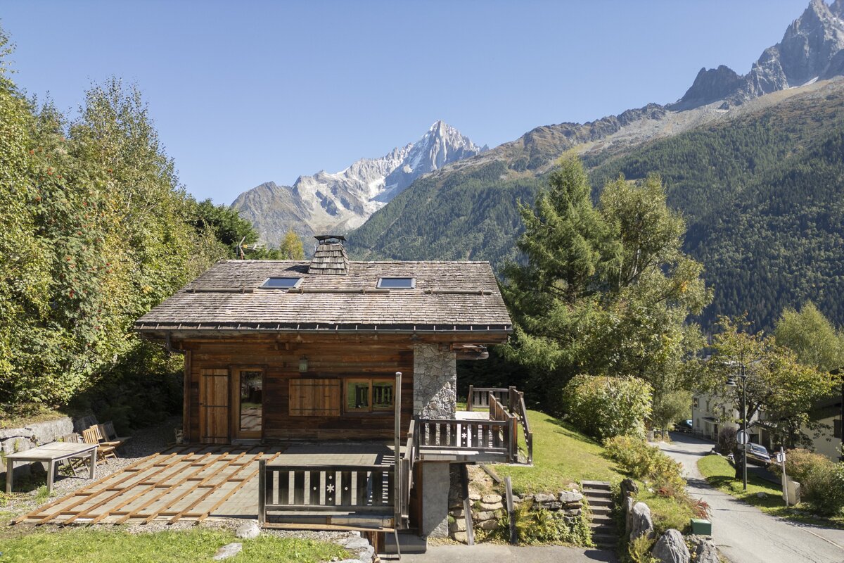 A rustic wooden chalet on a green hillside with majestic, snow-capped mountains in the background under a clear blue sky.